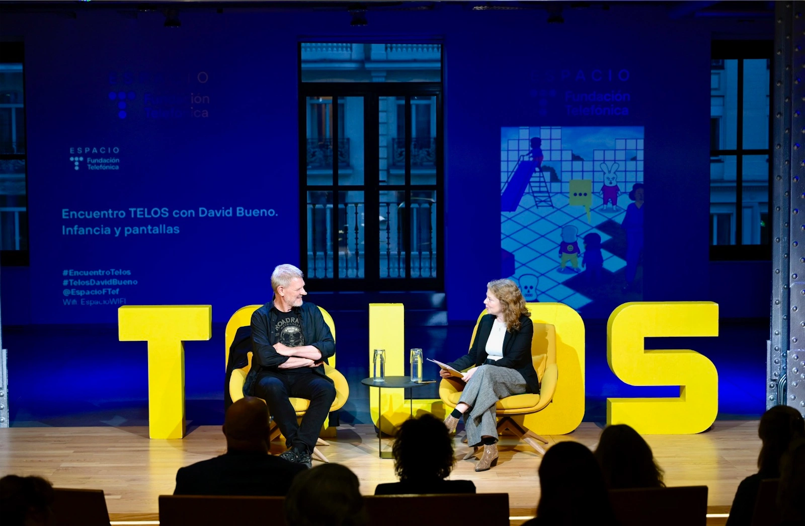 David Bueno y Eva Catalán conversan sentados en sillones amarillos sobre un escenario iluminado, con letras grandes que forman la palabra 