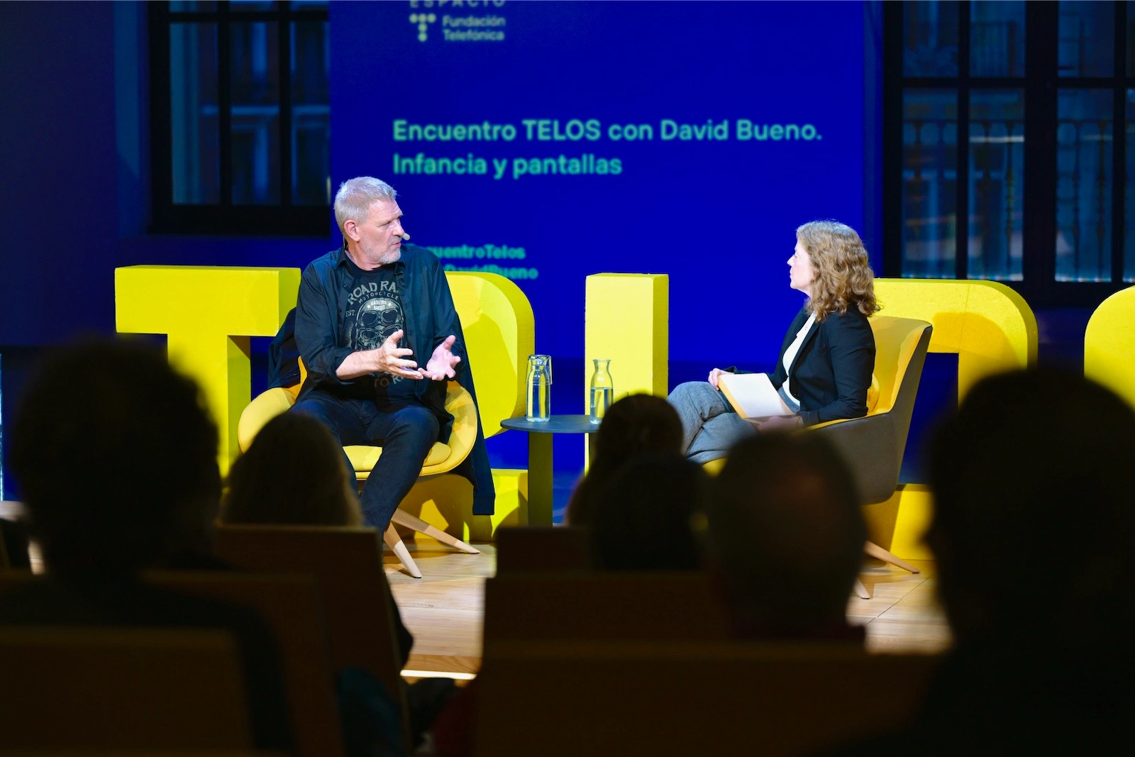 David Bueno y Eva Catalán dialogan en el escenario del auditorio Espacio Fundación Telefónica, sentados frente al público, con botellas de agua sobre una mesa entre ellos y letras gigantes de 